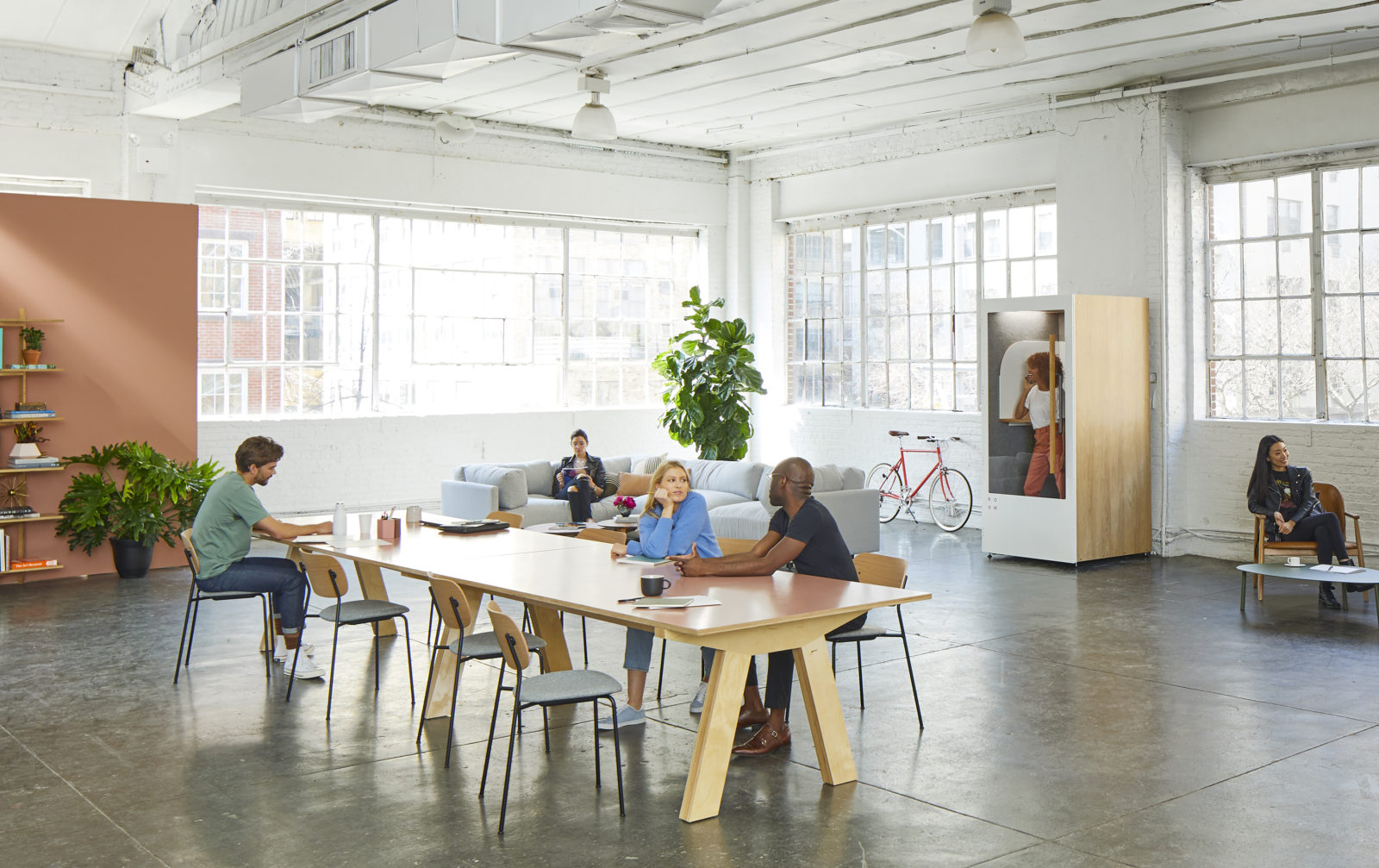Spacious, modern office with large windows, people working at a long table, one person on a sofa, and another sitting near a bookshelf with plants and a red bicycle.