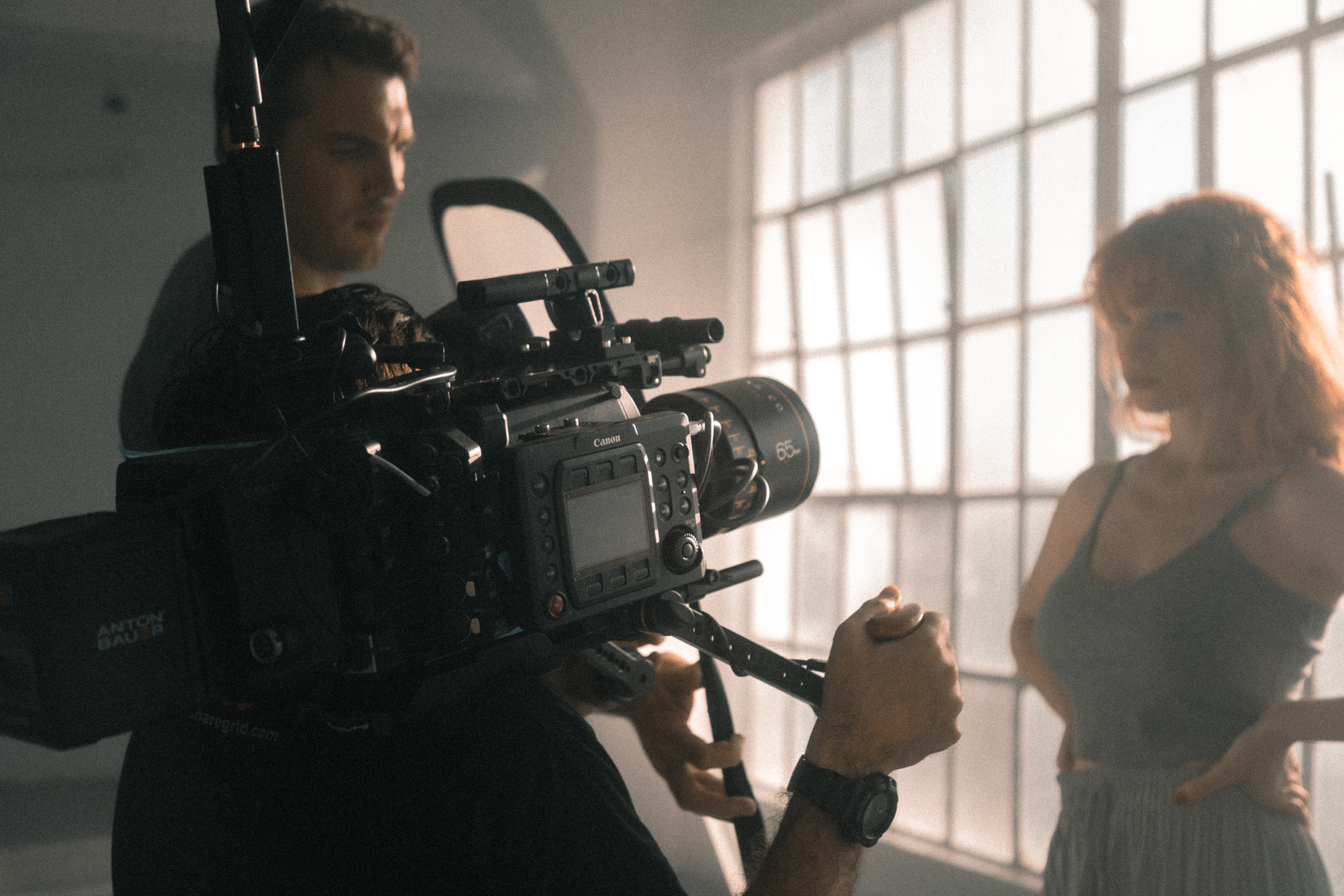 A film crew records a woman standing near large windows in a well-lit room, with a professional video camera in the foreground.