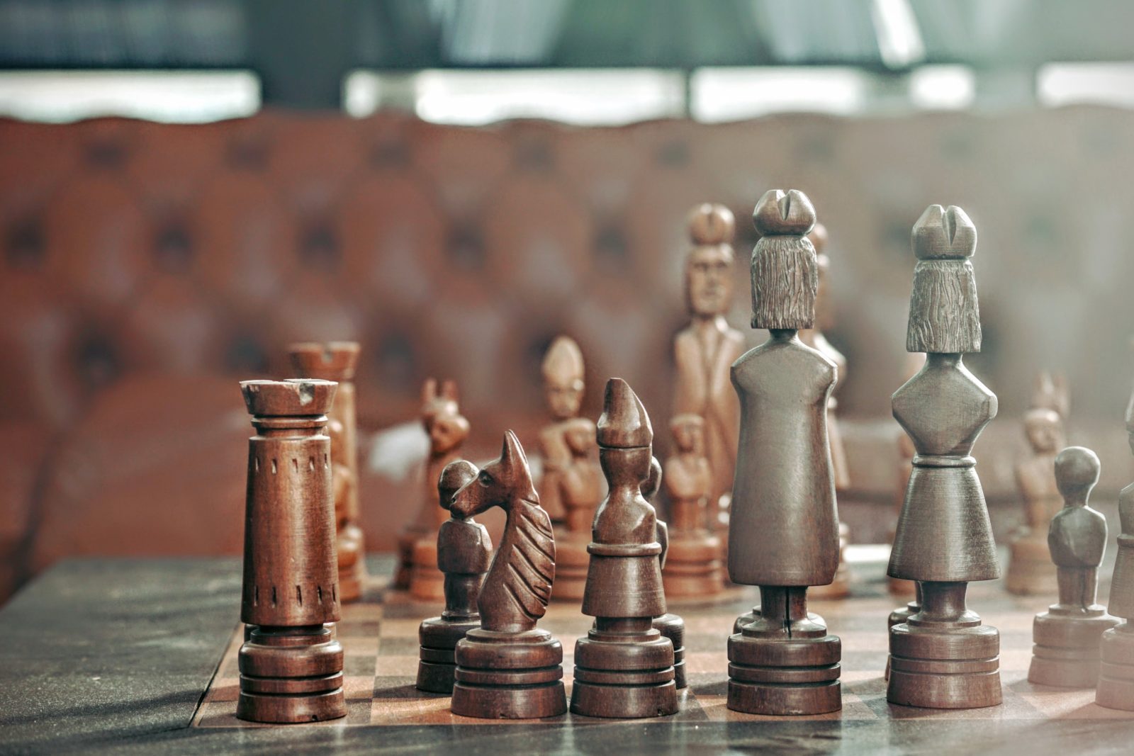 Close-up of a wooden chessboard with uniquely carved chess pieces, focused on the white side, against a blurred background.