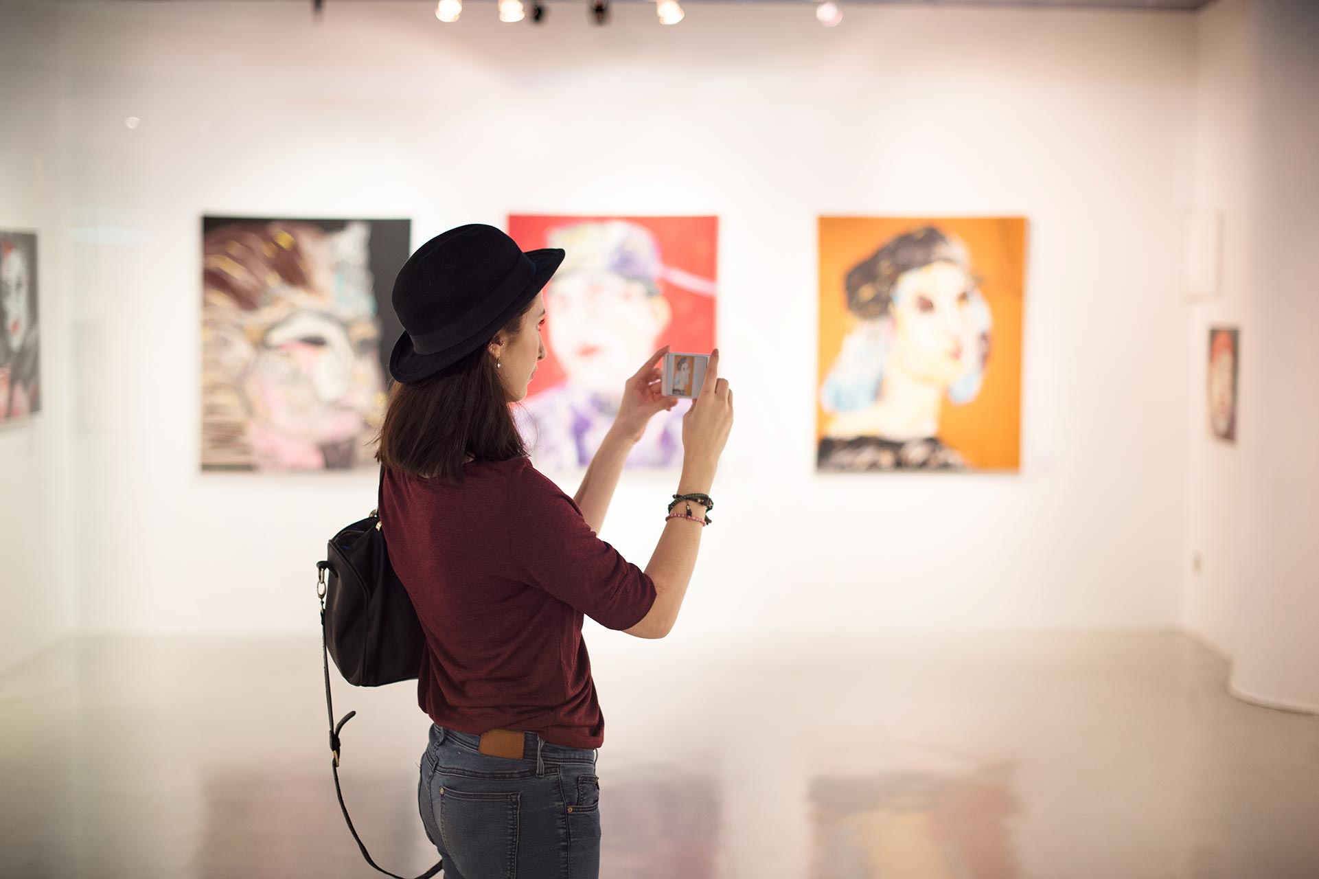 A woman wearing a black hat and red shirt takes a photo with her phone in an art gallery, mindful of art law and licensing, as three colorful portraits hang on the wall.
