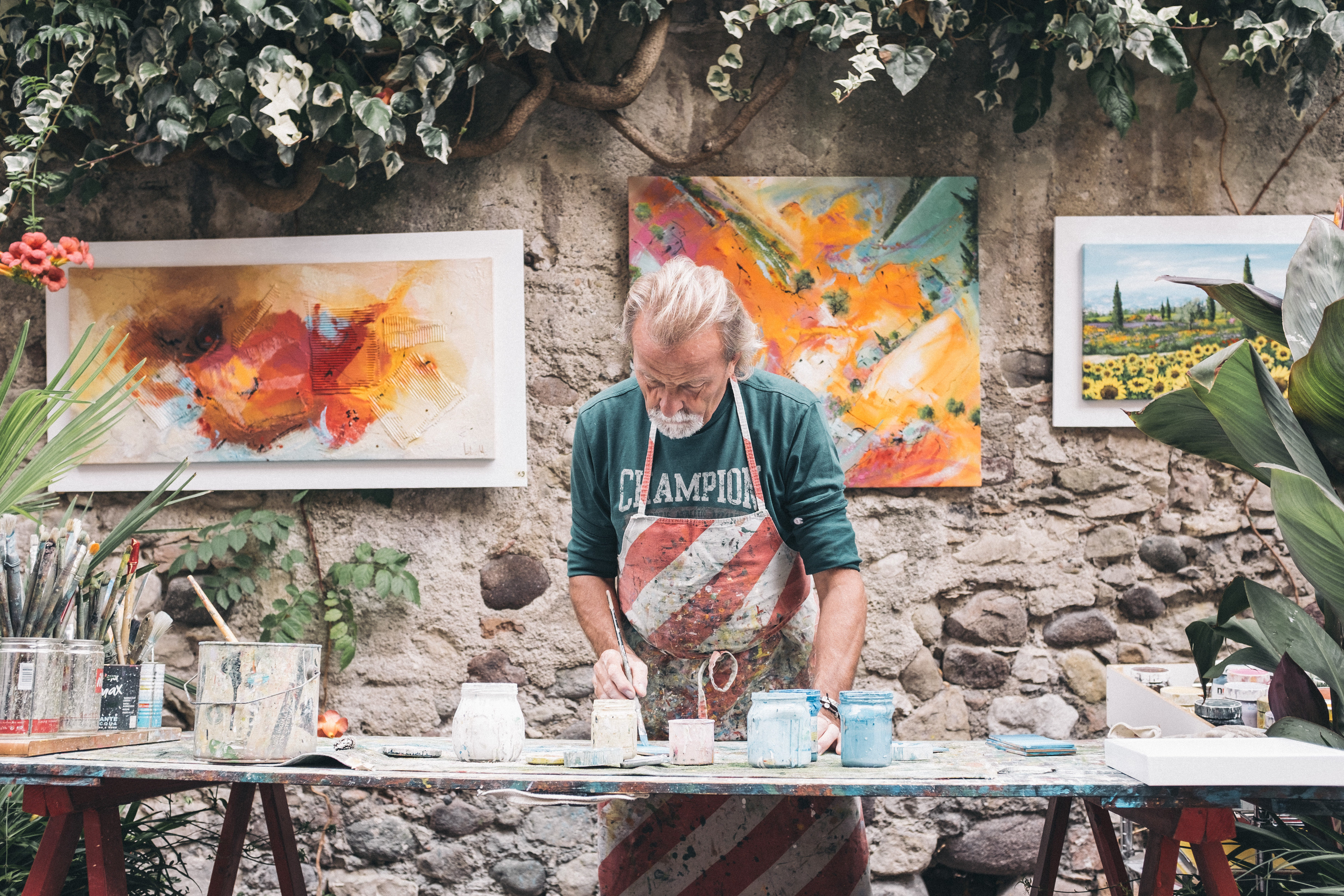 An older man wearing an apron paints at an outdoor table covered in art supplies, with colorful abstract and landscape paintings hanging on a stone wall behind him.