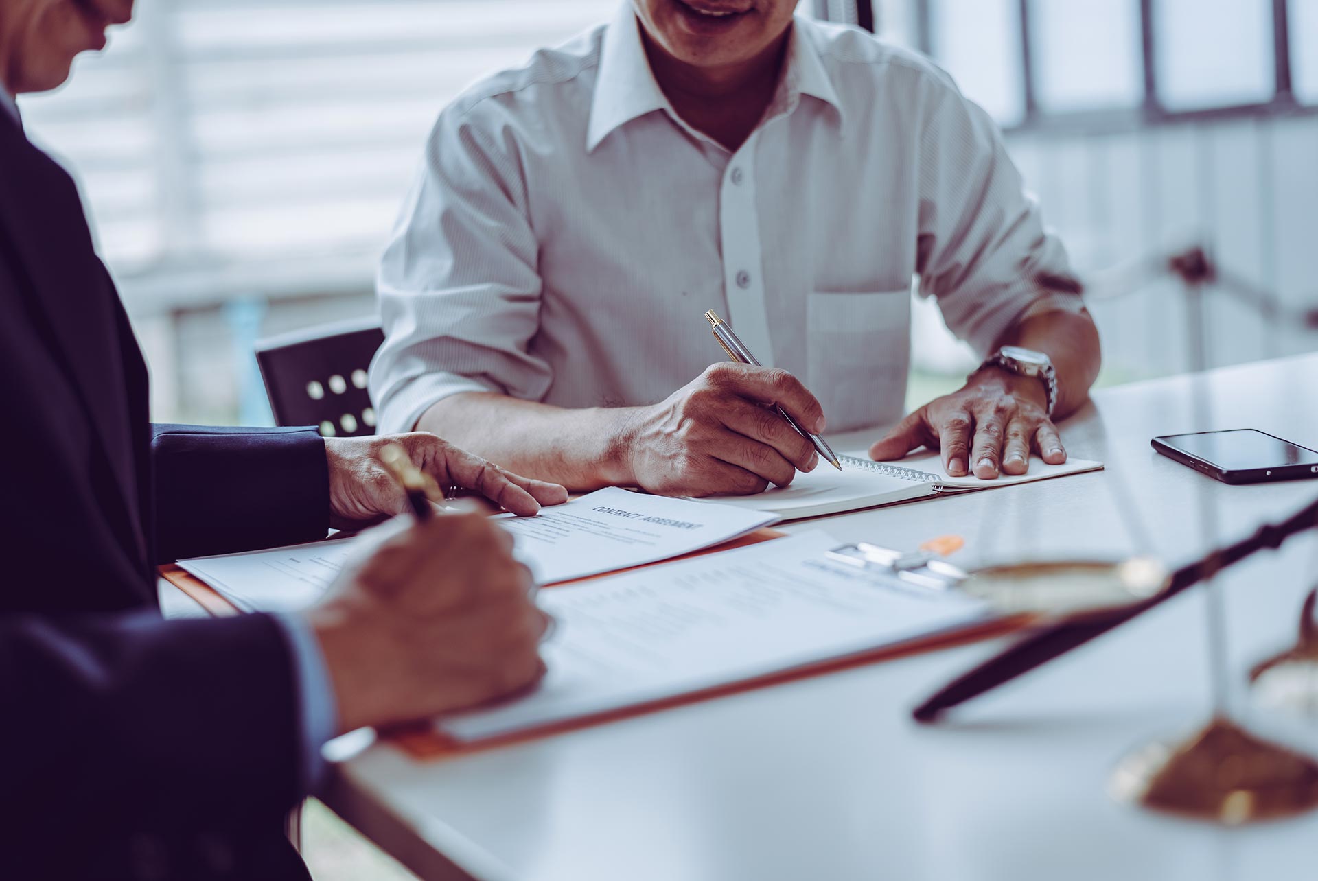 Two people sit at a desk signing business contracts, with pens in hand and papers in front of them; a smartphone lies on the table.