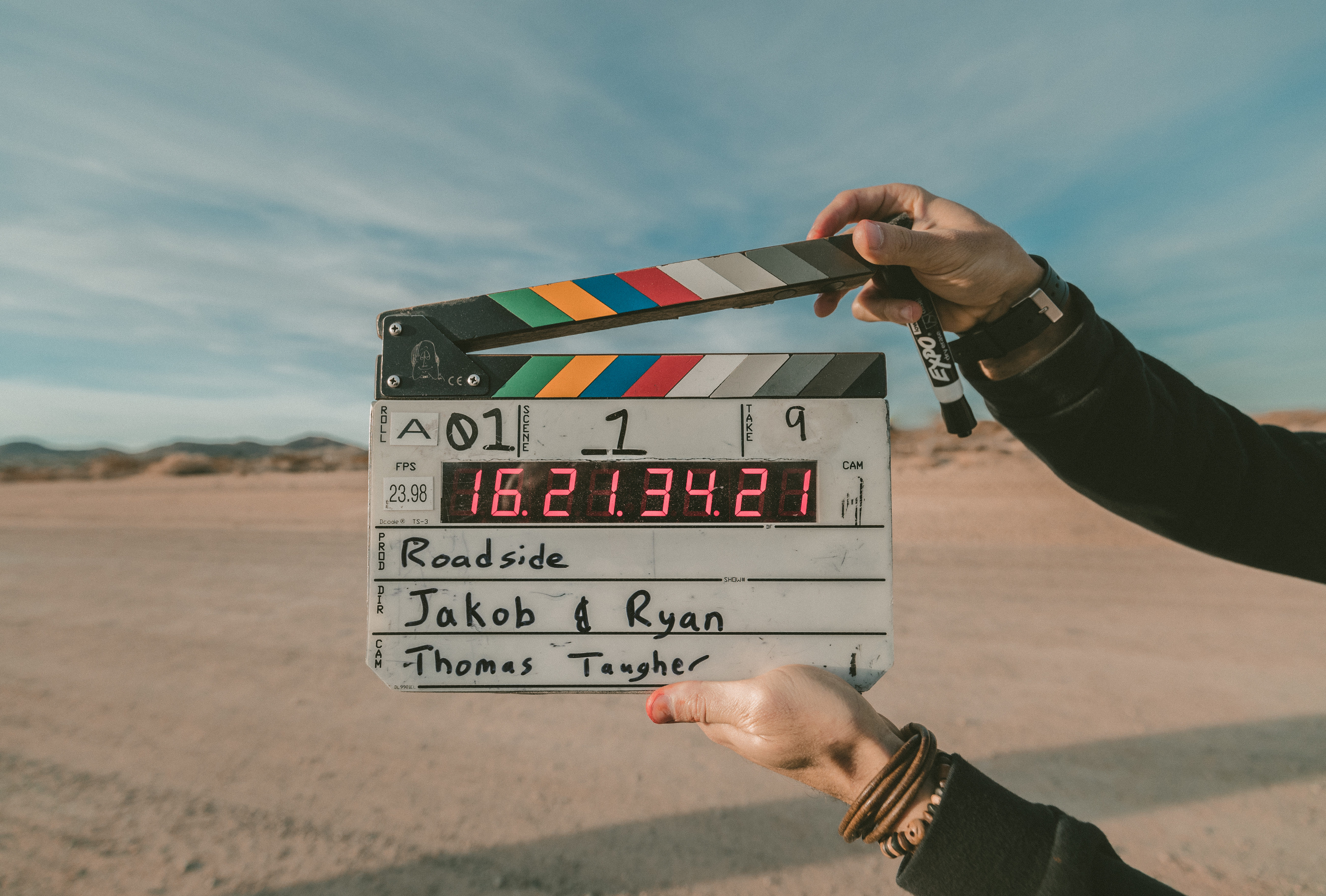 A person holds a film clapperboard outdoors in a desert setting. The clapperboard reads "Roadside," with names Jakob & Ryan and Thomas Taugher written on it.