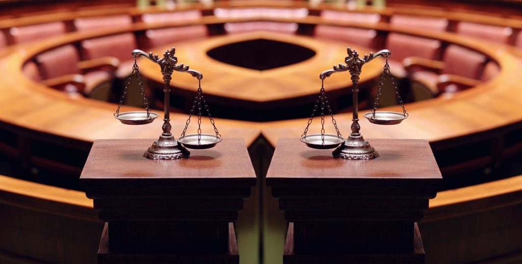 Two scales of justice placed on wooden podiums in an empty courtroom with curved rows of seats in the background, reflecting the environment where a commercial litigation attorney NYC might advocate for clients.
