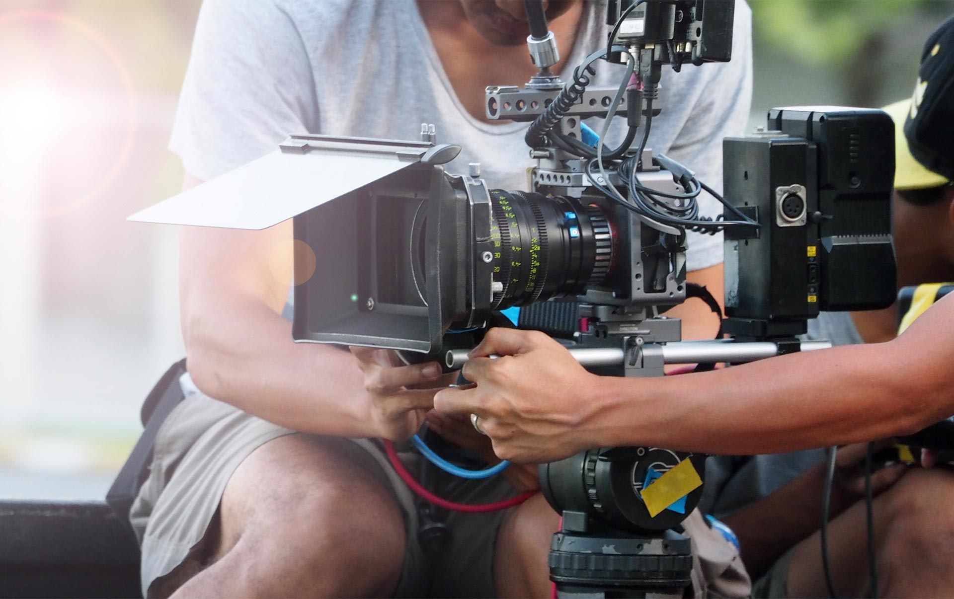 Two people adjust a professional video camera mounted on a tripod, preparing for an outdoor film shoot.