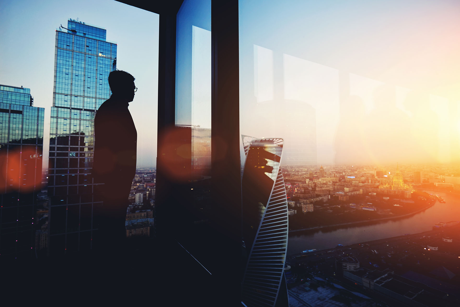 A person stands by a large window in a high-rise building, overlooking a cityscape at sunset with tall glass buildings and a river visible.