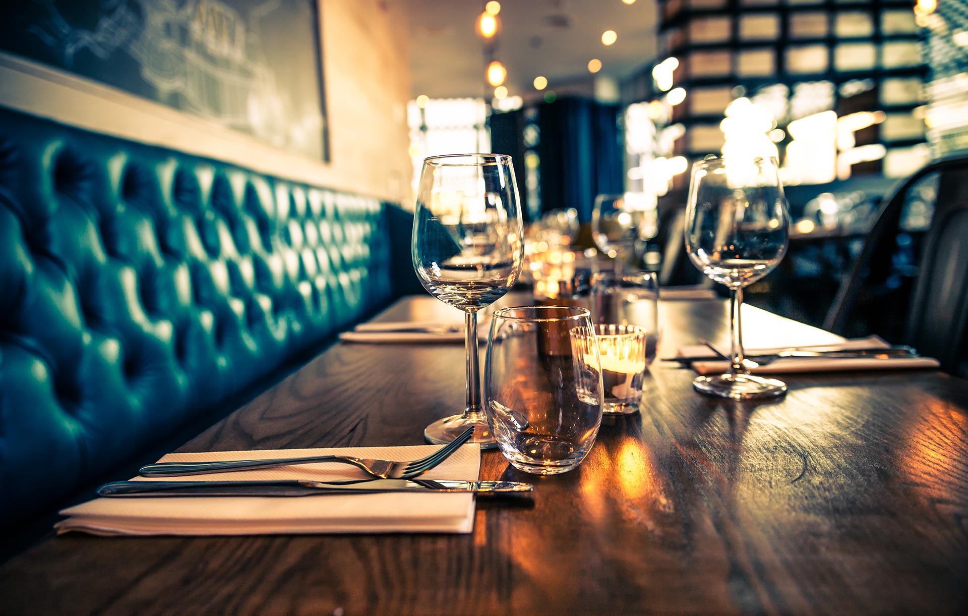 A restaurant table set with glasses, cutlery, and napkins, featuring teal upholstered seating and softly lit candles in the background—perfect for an evening out at a venue with full alcohol licensing.