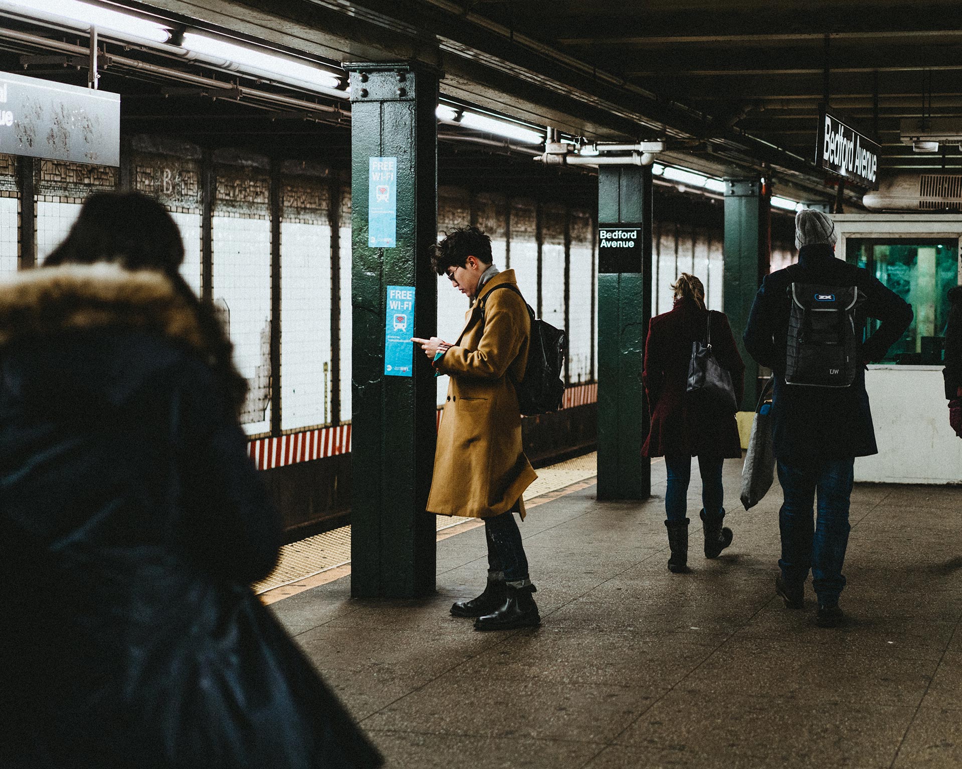People wait on a subway platform; one person in a tan coat stands near a column looking at their phone, while others walk by. The station sign reads Bedford Avenue.