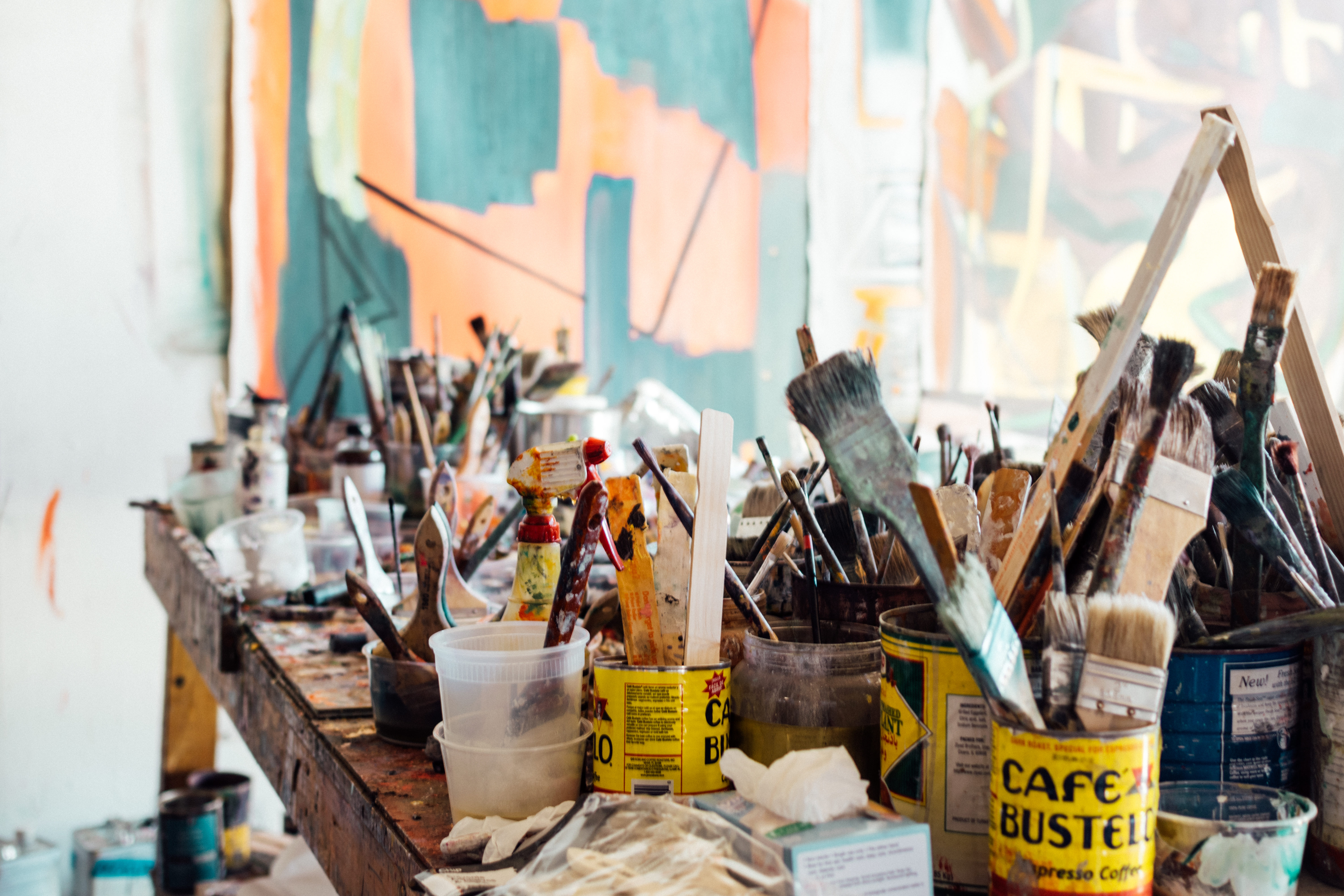 A cluttered artist's workspace with paintbrushes, paint cans, and art supplies on a wooden table; abstract paintings are visible in the background.