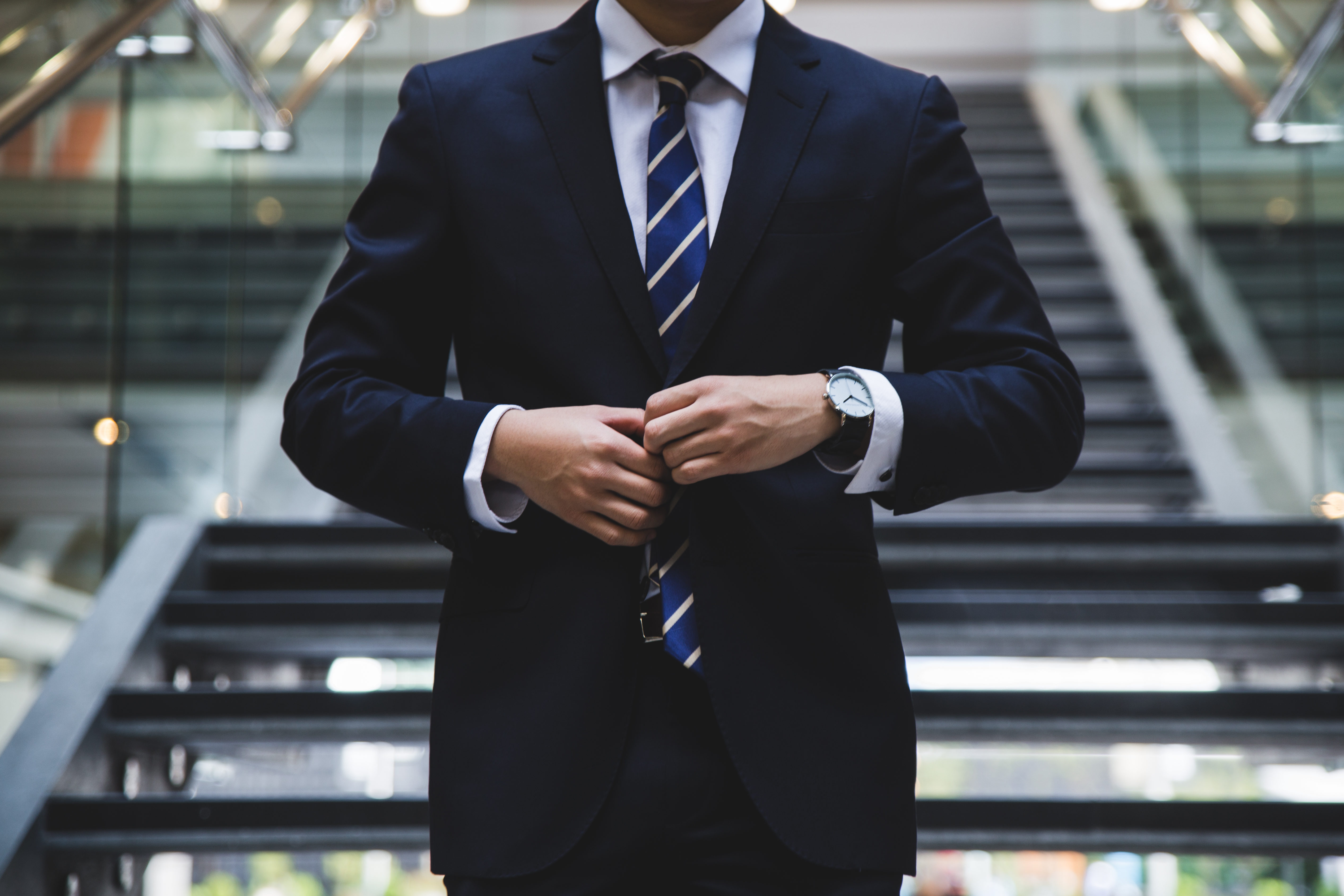 Man in a dark suit and striped tie adjusting his jacket while standing in front of stairs indoors.