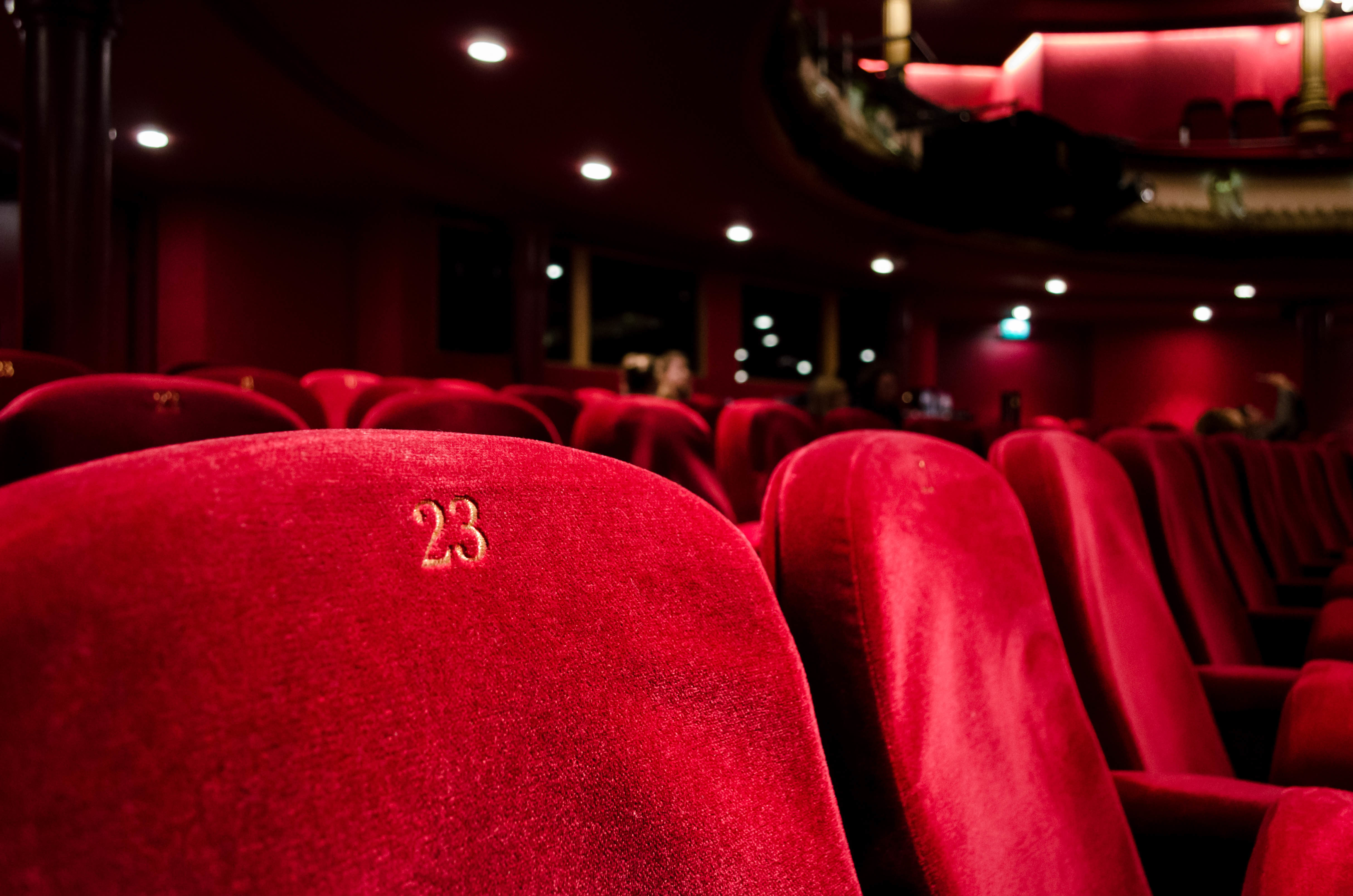 Close-up of red velvet theater seats, with seat number 23 in focus, in a dimly lit auditorium.