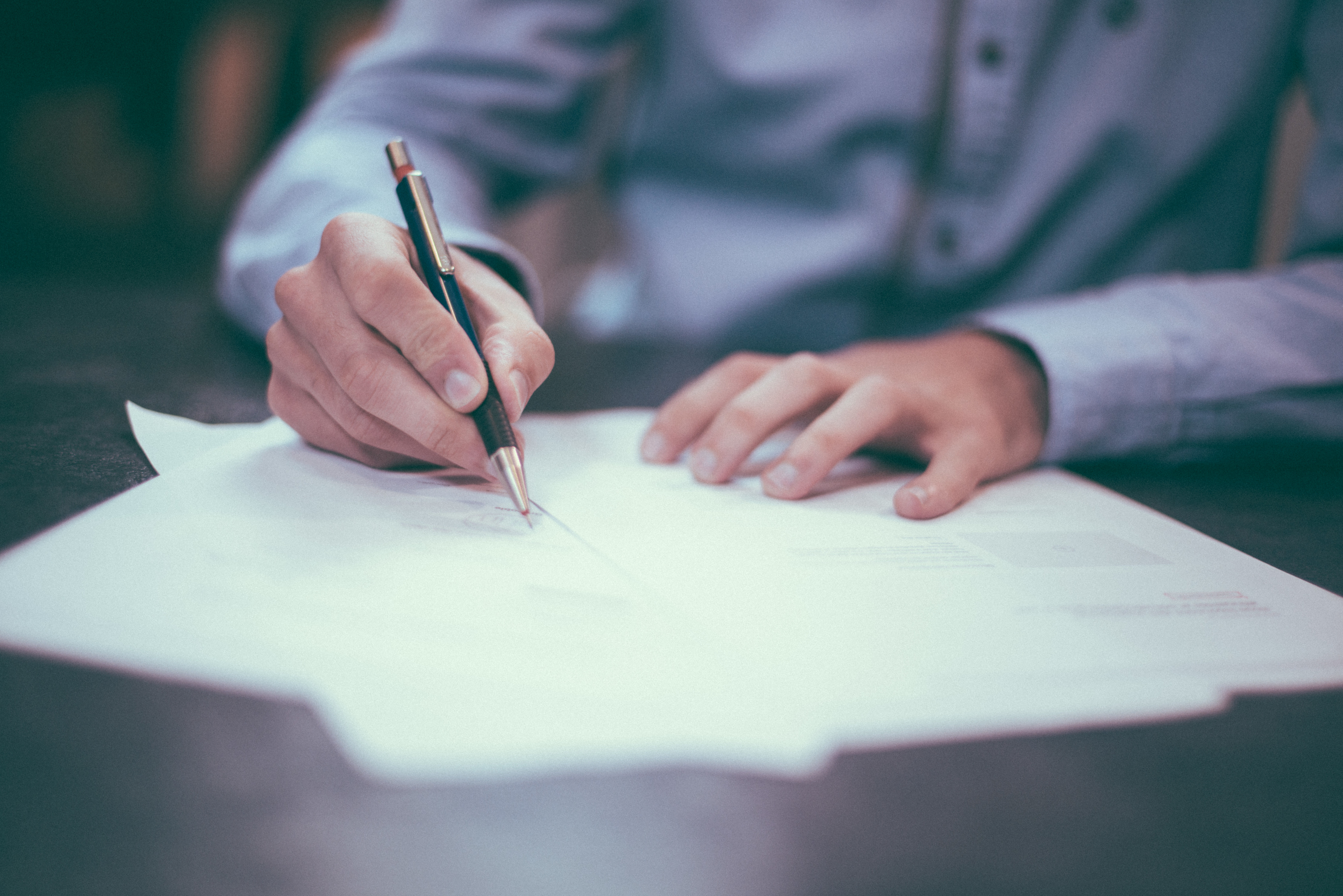 A person in a button-up shirt writes on paper documents with a pen at a desk.