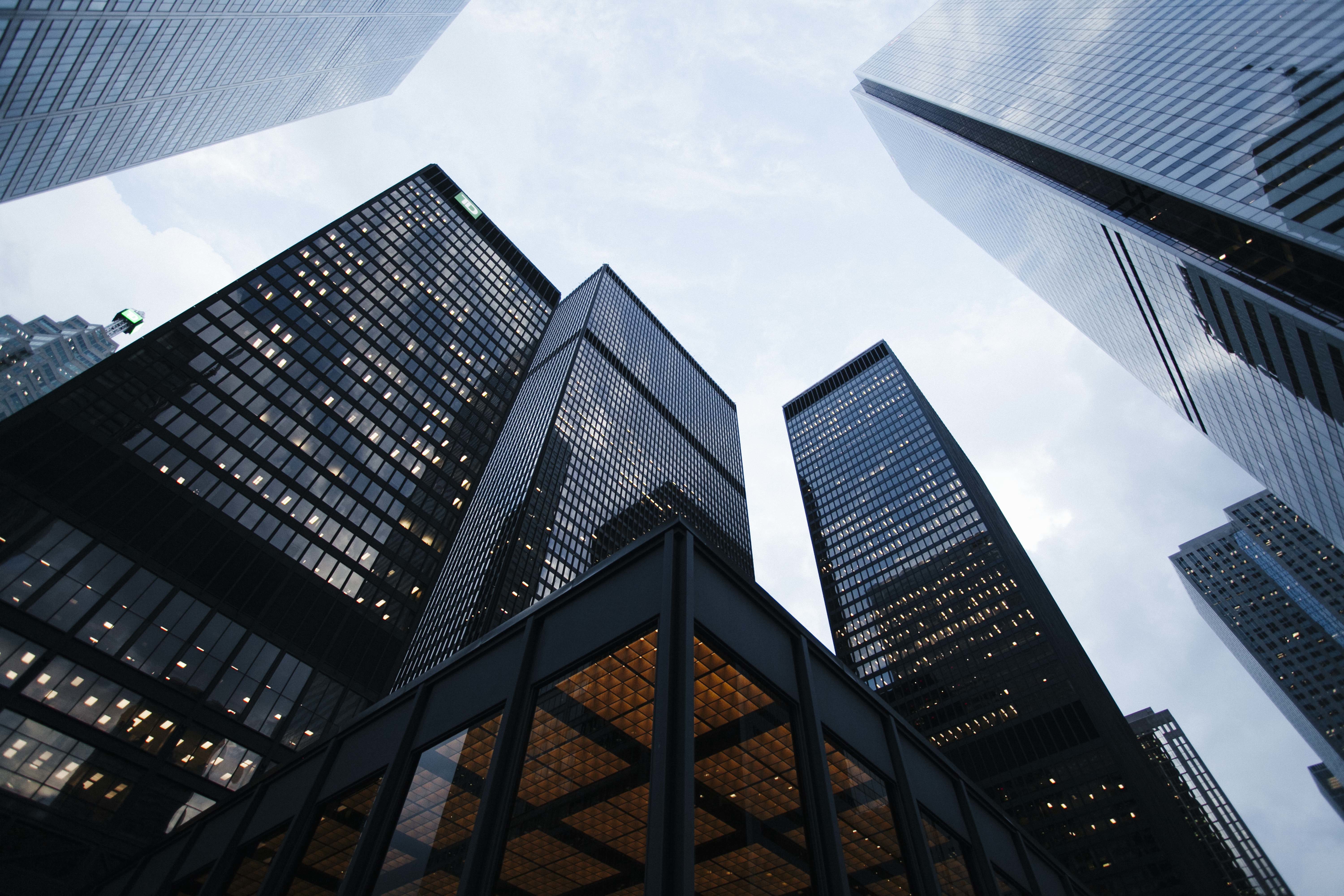 Tall modern skyscrapers viewed from below against a cloudy sky, with glass exteriors reflecting light and nearby buildings.
