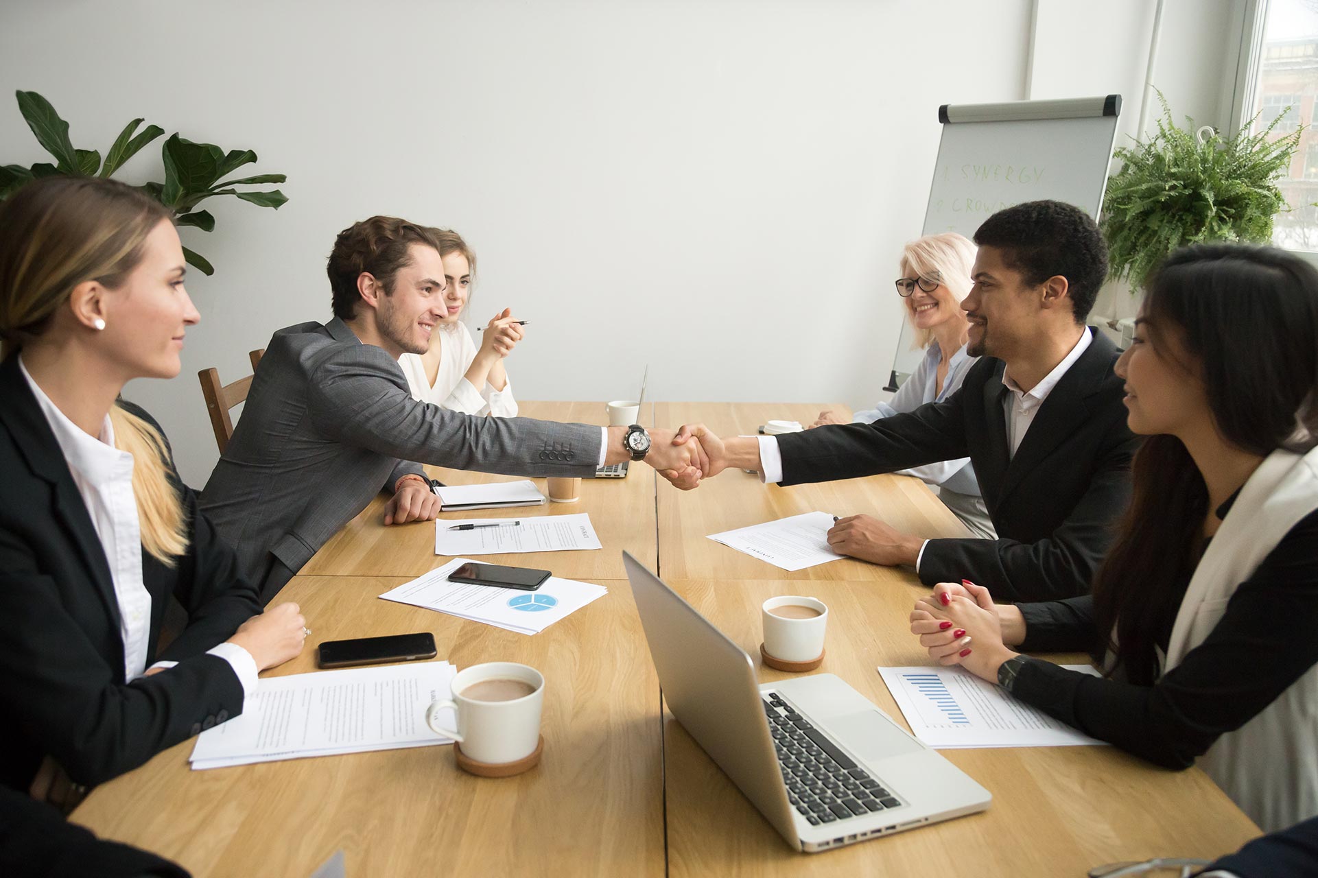 Two men shaking hands during a business meeting with several others seated around a table