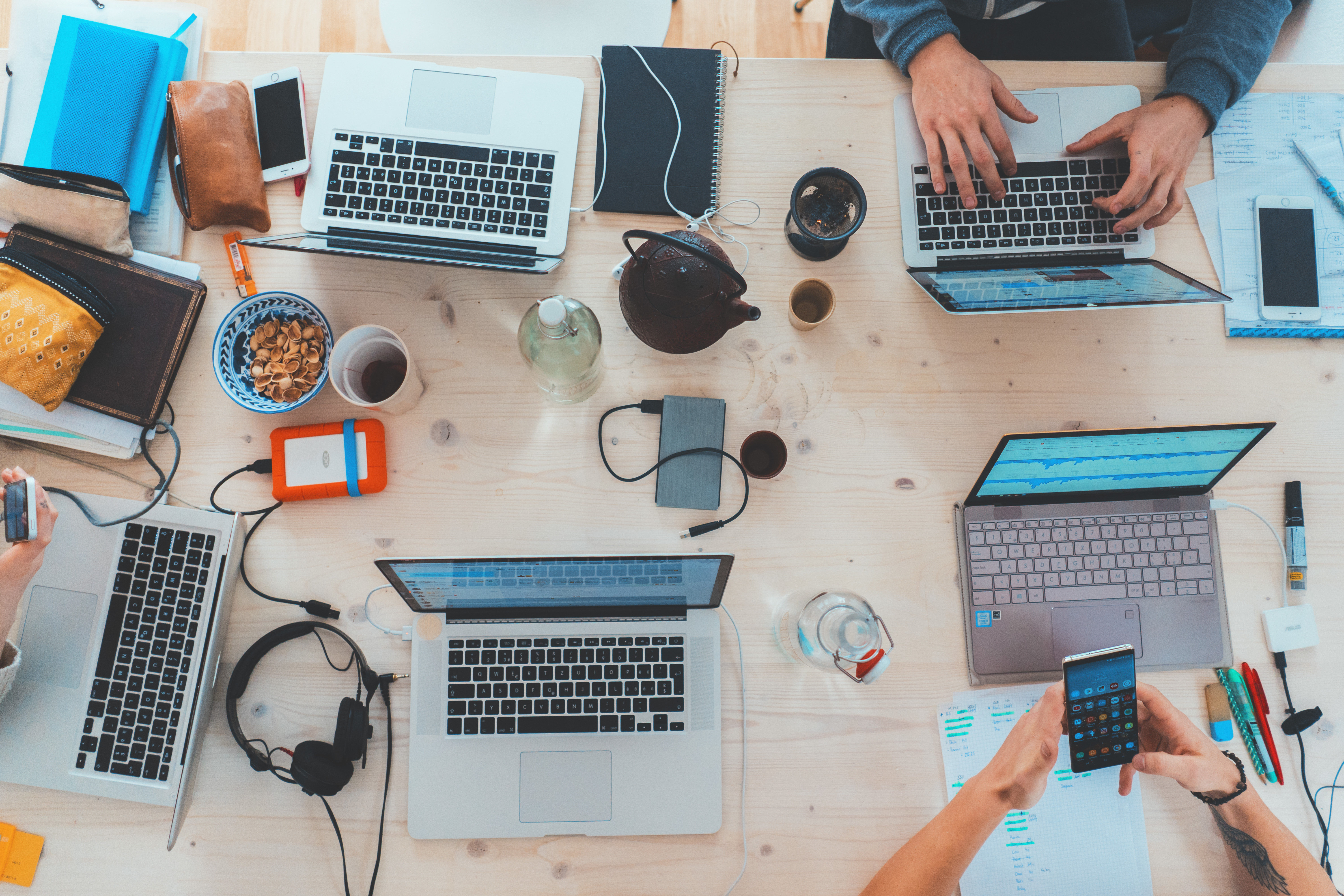 Overhead view of a wooden table with several laptops, notebooks, smartphones, snacks, and drinks, with four people working collaboratively.