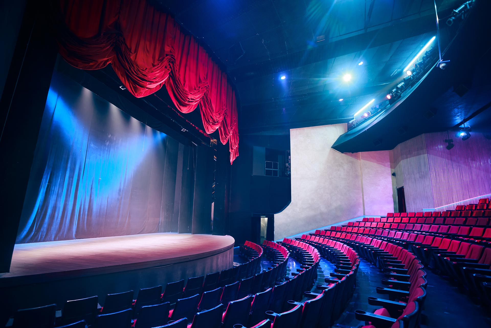 A quiet theater with rows of red seats faces the stage, where drawn curtains glow under dim blue and purple lighting.