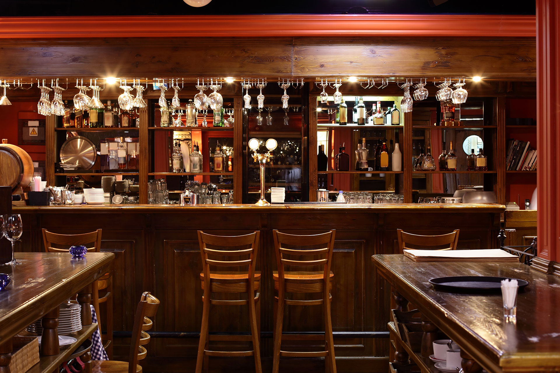 A wooden bar with high chairs, shelves stocked with bottles and glassware, and wine glasses hanging above the counter in a warmly lit restaurant free from violations and boasting top grading.