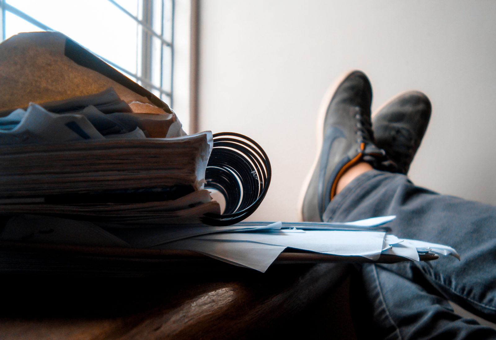 A person with feet up on a desk wearing sneakers sits next to a stack of disorganized papers and folders near a window.