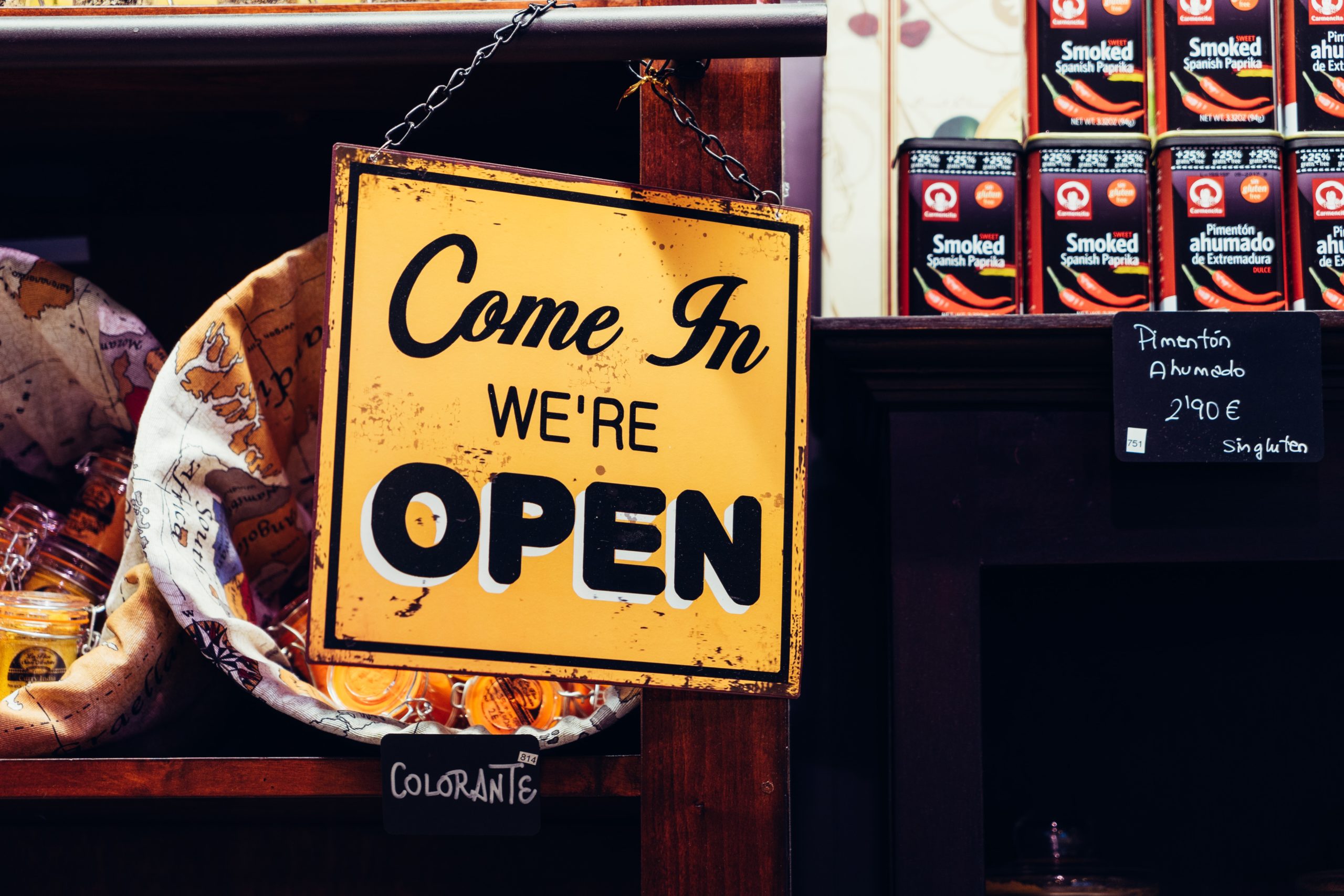 Yellow "Come In, We're Open" sign hanging in a store with shelves displaying various packaged goods in the background—a welcoming scene full of Small Business Lessons.