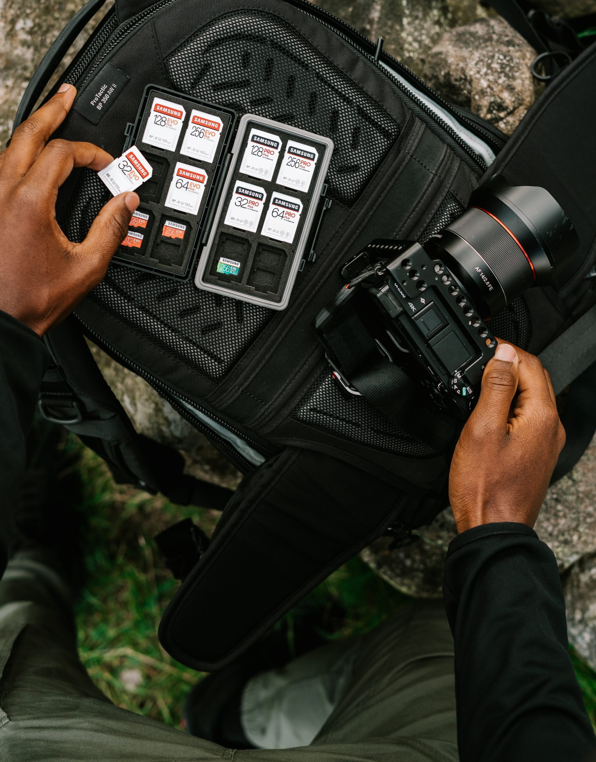 A person prepares a camera and organizes multiple memory cards on an open camera backpack outdoors.