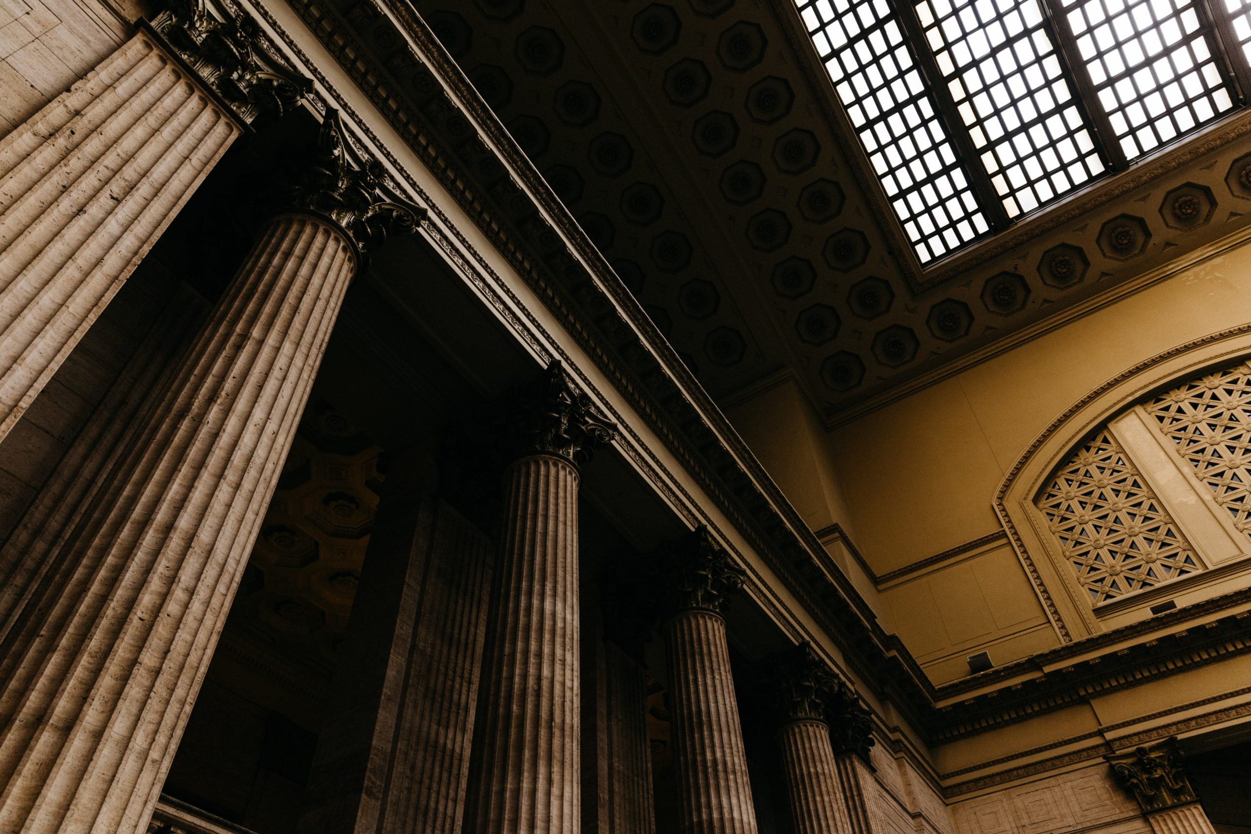 Interior view of a grand building with tall classical columns and detailed ceiling, lit by large grid-patterned skylights.