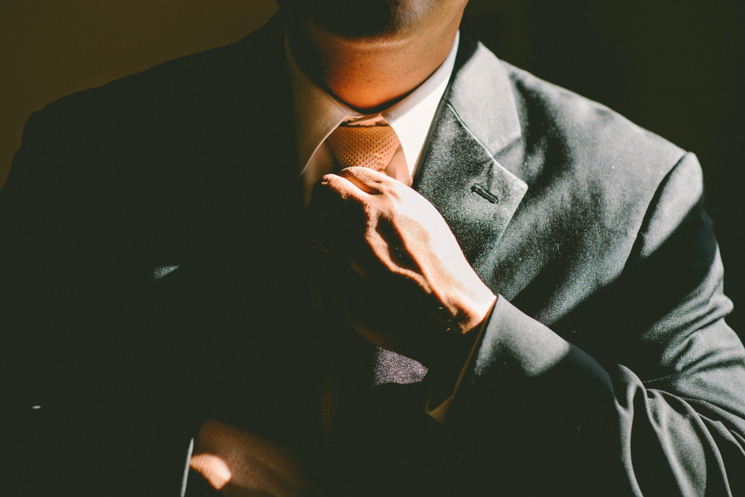 A person in a suit adjusts their tie with one hand, with their face partially obscured by shadow and sunlight.