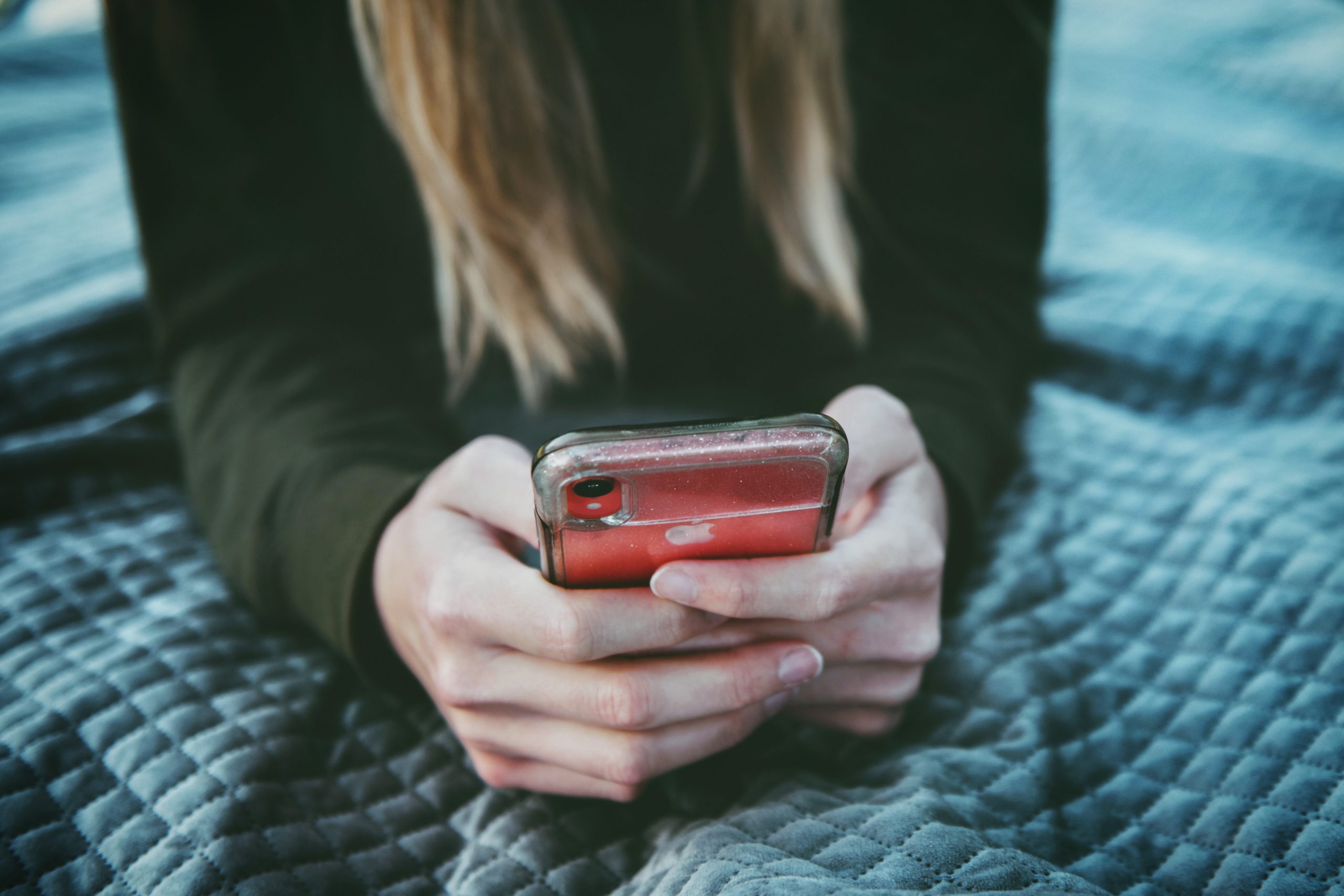 Person with long, light brown hair holding a red smartphone while lying on a quilted blue blanket.