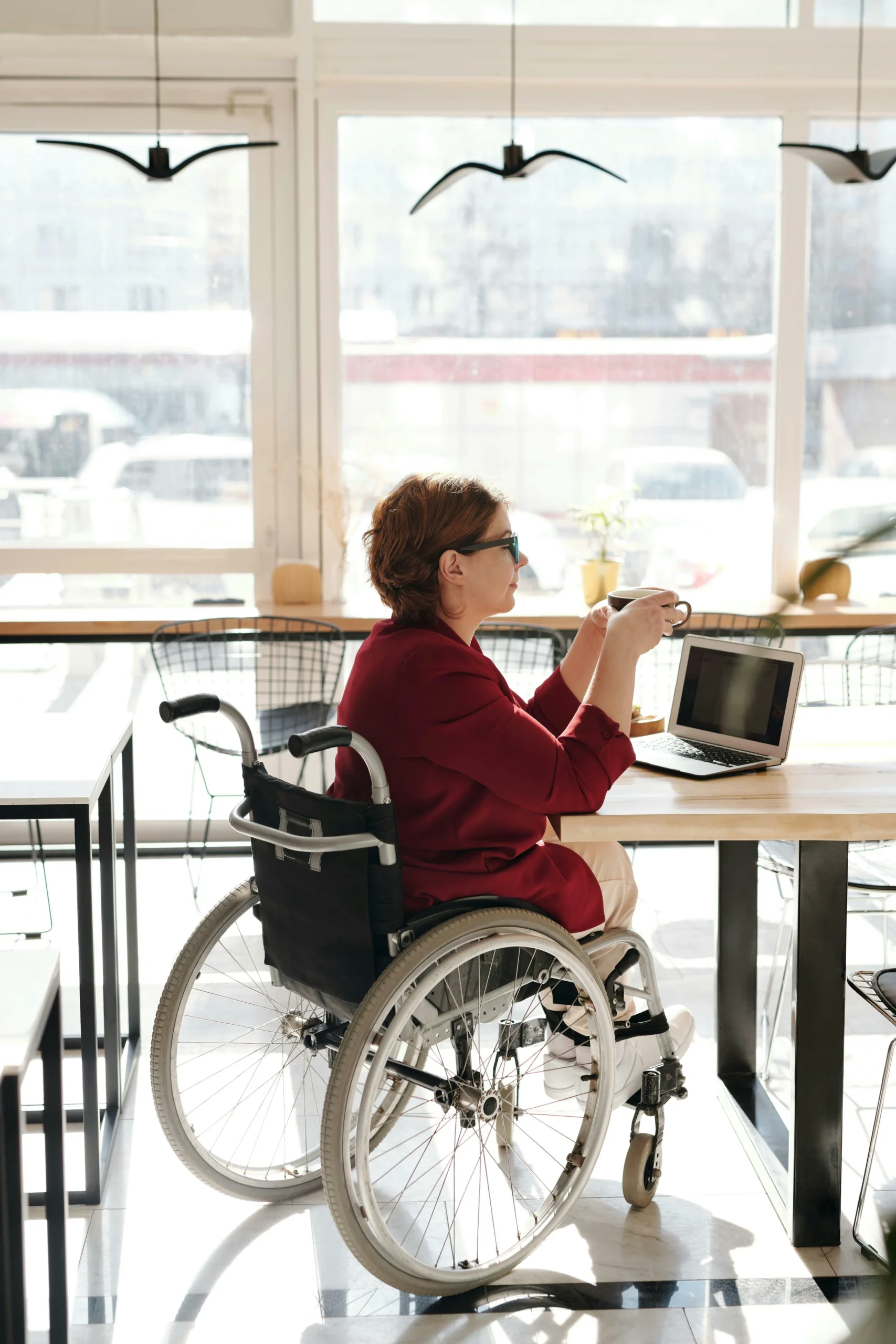 A woman in a wheelchair sits at a table in a bright café, holding a cup and working on a laptop.