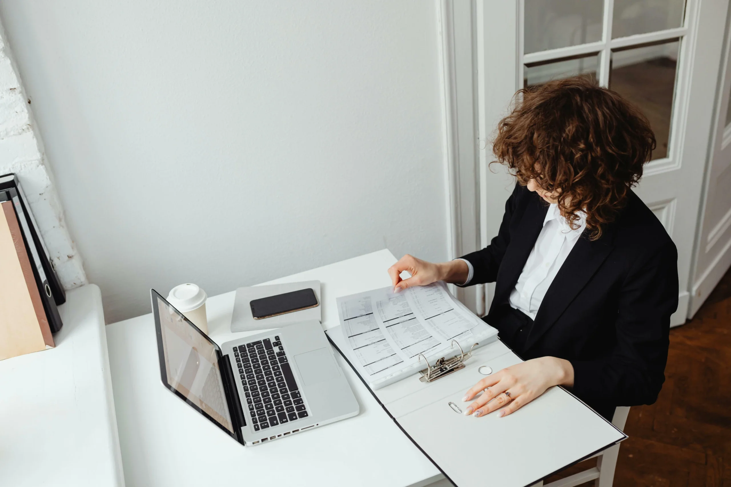 Person in business attire sits at a desk reviewing documents in a binder, with an open laptop, smartphone, and coffee cup nearby.