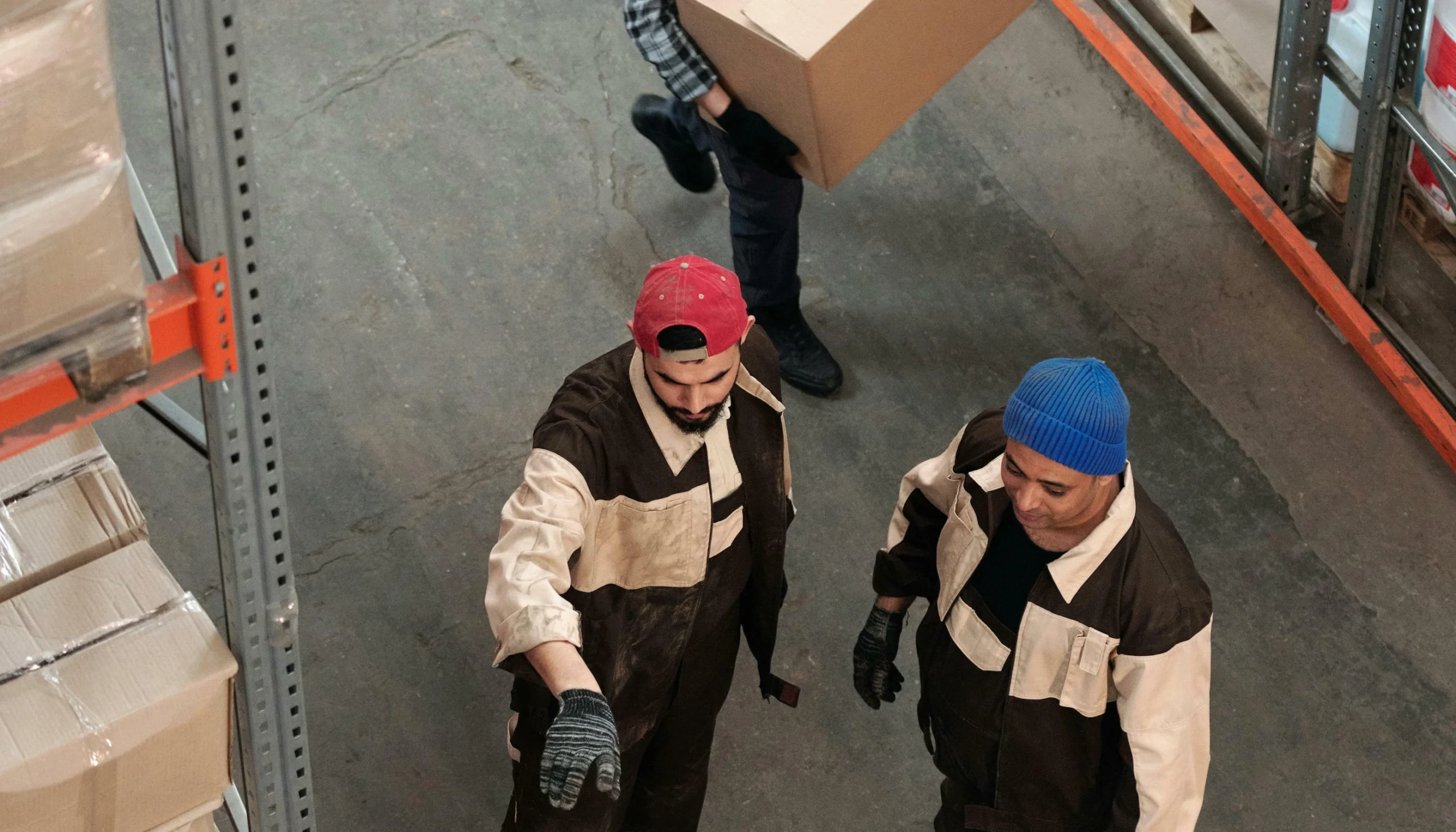 Two warehouse workers in uniforms and gloves stand on a concrete floor, one pointing ahead, while a third person carries a box in the background.