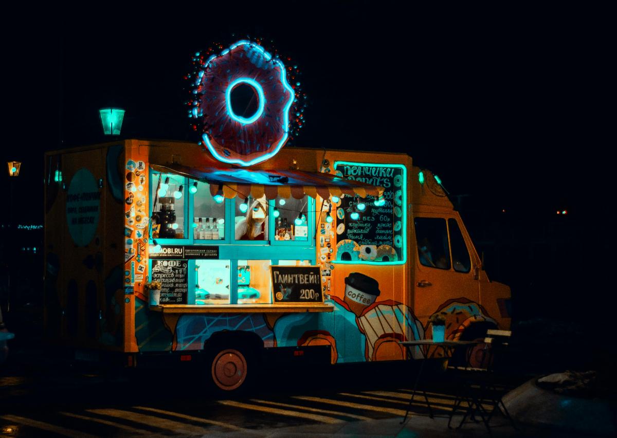 A brightly lit food truck with a large glowing donut sign on top, parked at night with no customers around.