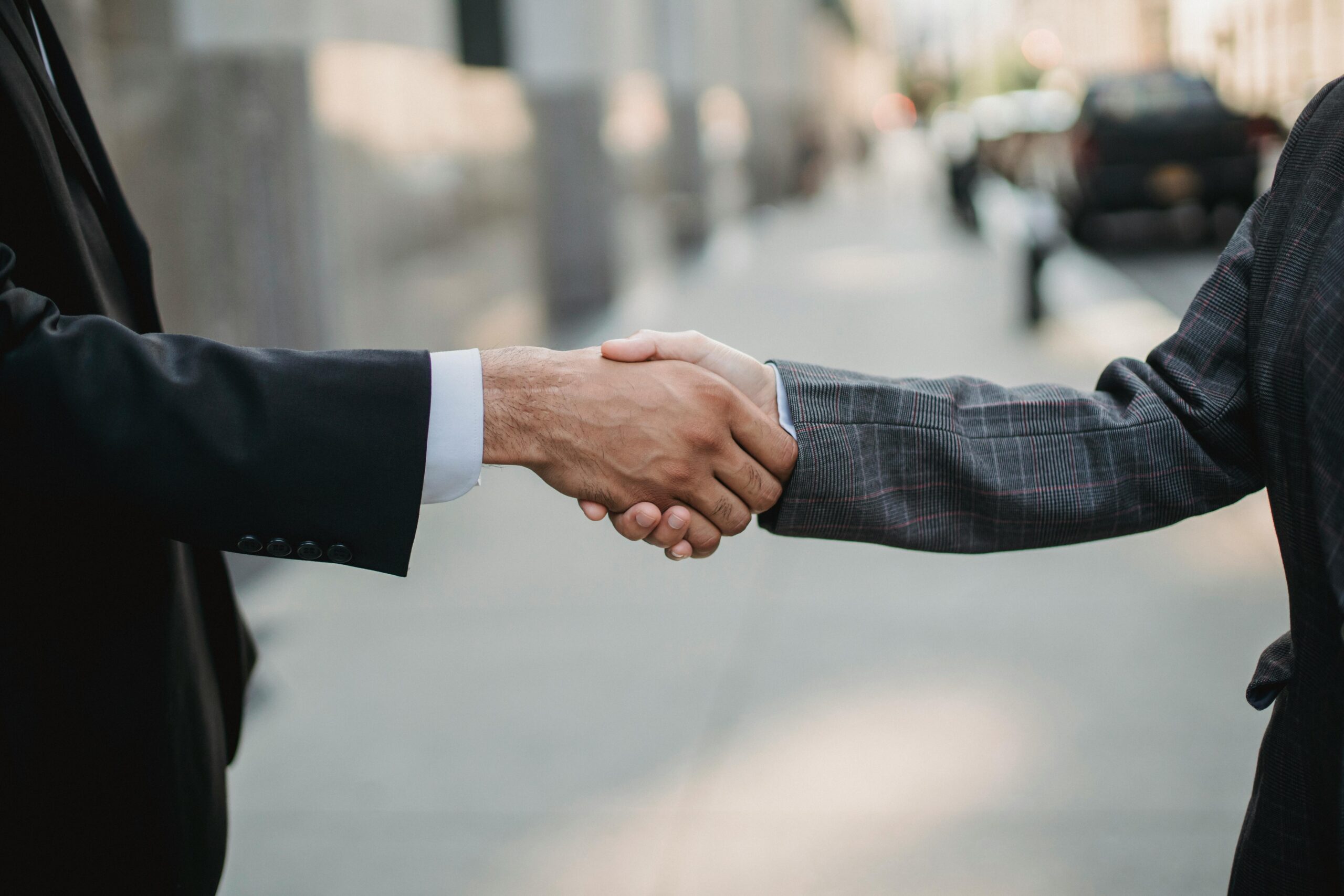 Two people wearing business suits shake hands on a city sidewalk.