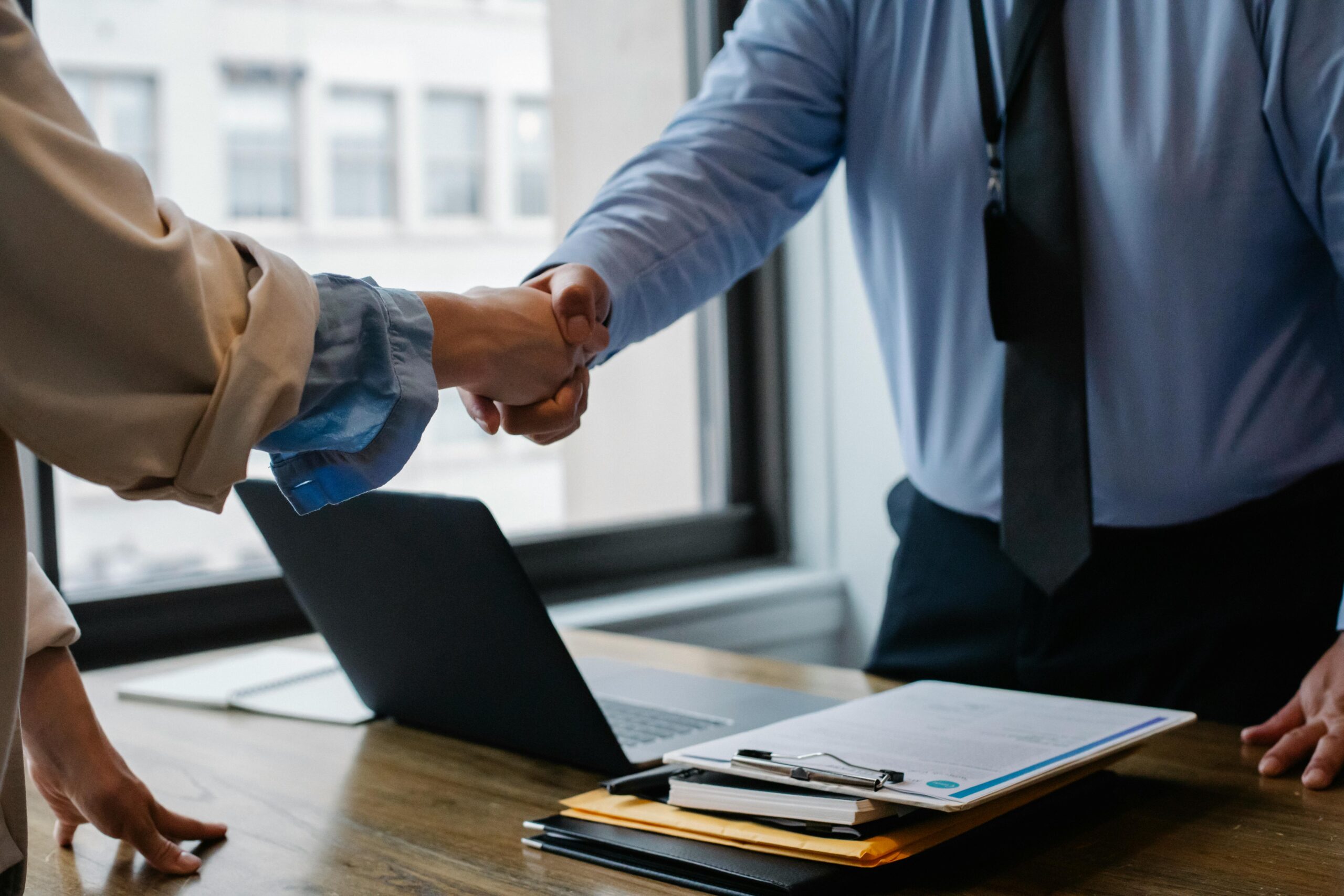 Two people shaking hands over a desk with papers, a clipboard, and a laptop in an office setting.