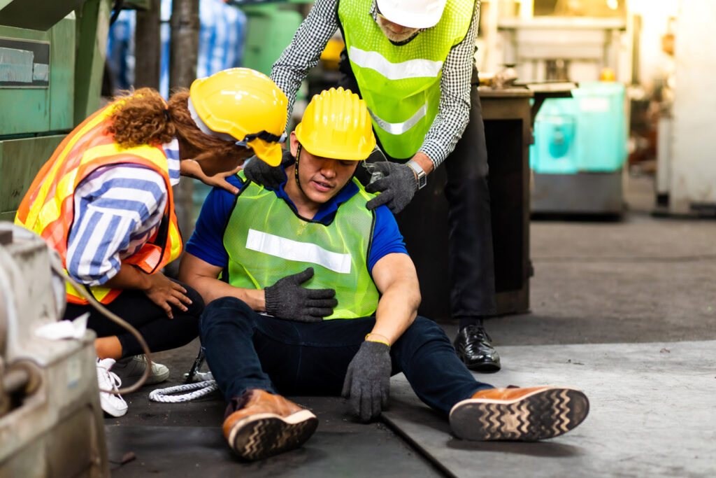 A construction worker sits on the floor appearing injured while two coworkers in safety gear assist him in an industrial setting—a reminder of when consulting a NYC personal injury lawyer may be necessary.