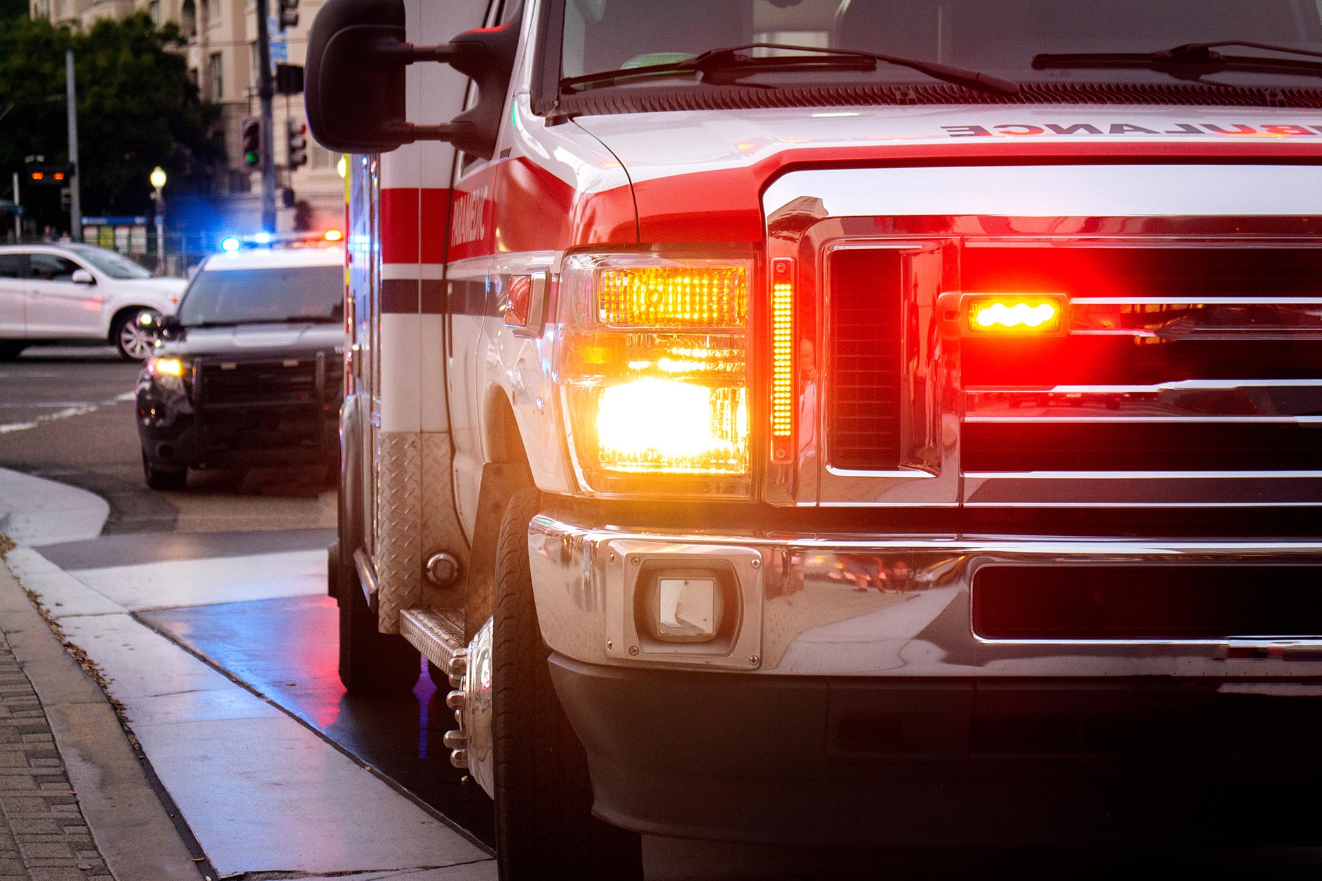 An ambulance with flashing lights is parked on a city street, as a police car and another vehicle—possibly belonging to business & litigation lawyers—are visible in the background.