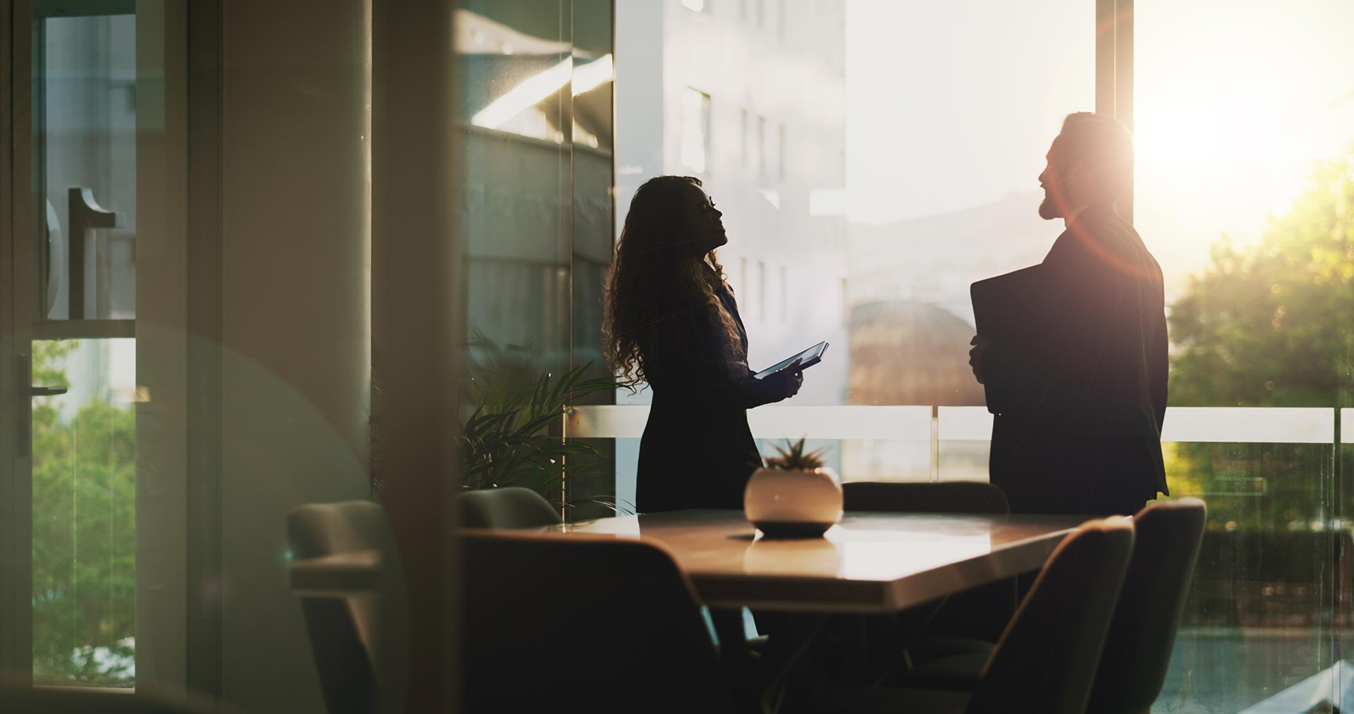 Two people stand and talk in an office by a large window with sunlight streaming in, one holding a tablet and the other a folder—business & litigation lawyers discussing important case details.